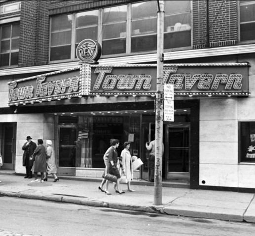 A black and white photograph of the Town Tavern's sign taken during the day with people walking by.