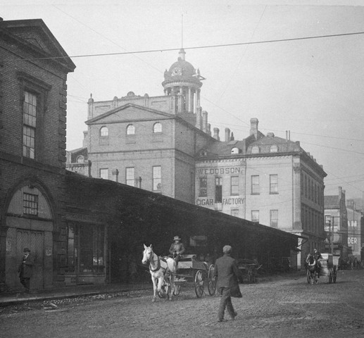 A black and white photograph of a street with pedestrians and horse drawn carriages walking along it. Buildings are visible to the left hand side of the image. You can see the Market in the foreground and St. Lawrence Hall in the back.