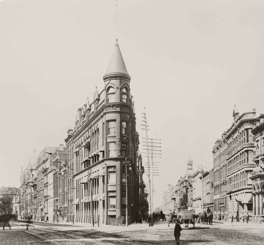 A black and white picture of the wedge-shaped Gooderham "Flatiron" Building at Wellington and Church Streets. A young boy is looking at the camera in the foreground. Several horse-drawn carts are on the street.