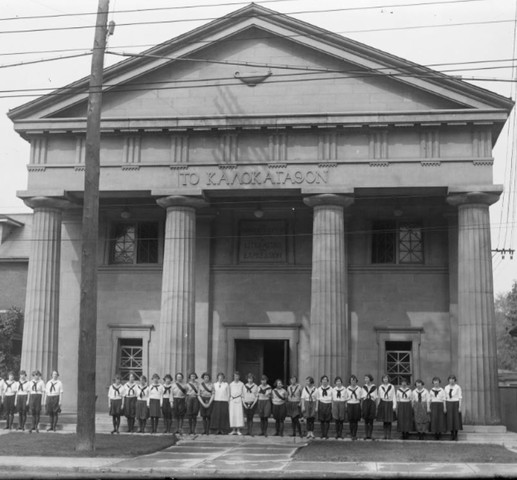 An archival photo shows a line of women in uniforms consisting of dark leggings, knee or ankle length skirts and neckerchiefs. They are standing in front of a building with four large columns that are two stories high.
