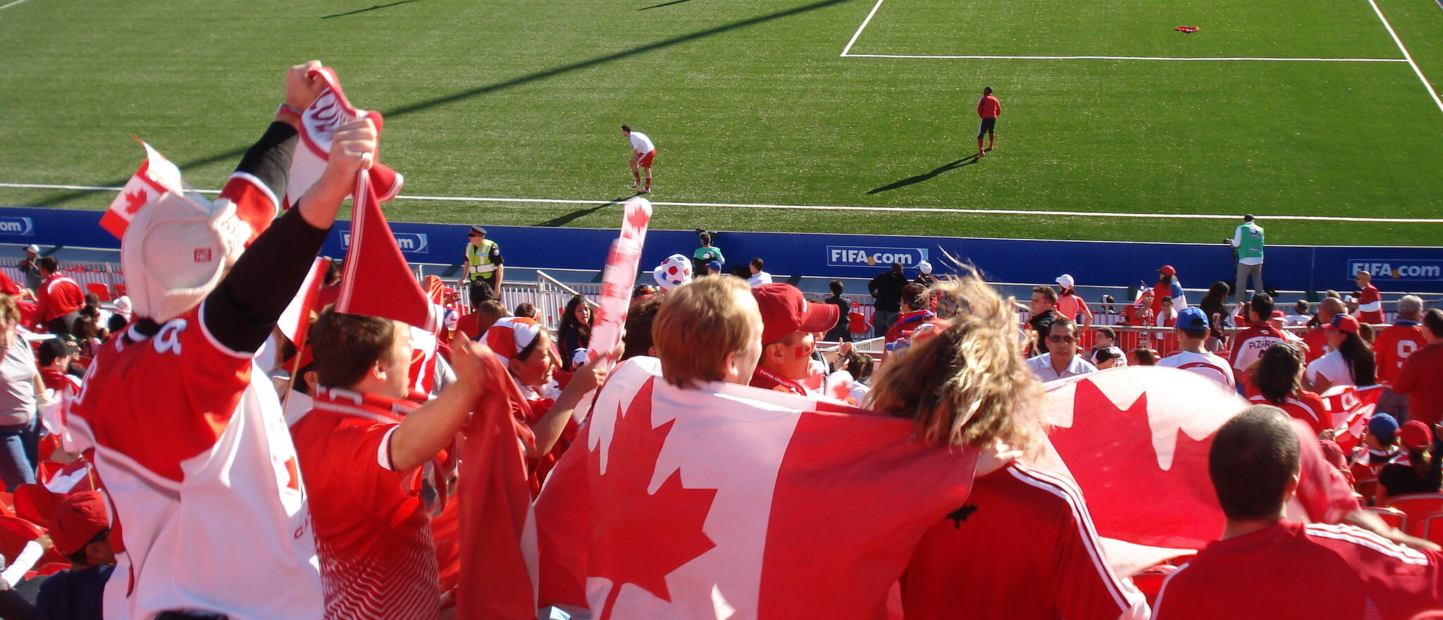 A large crowd of people wearing red and white and waving the flag of Canada, cheering at a soccer stadium.