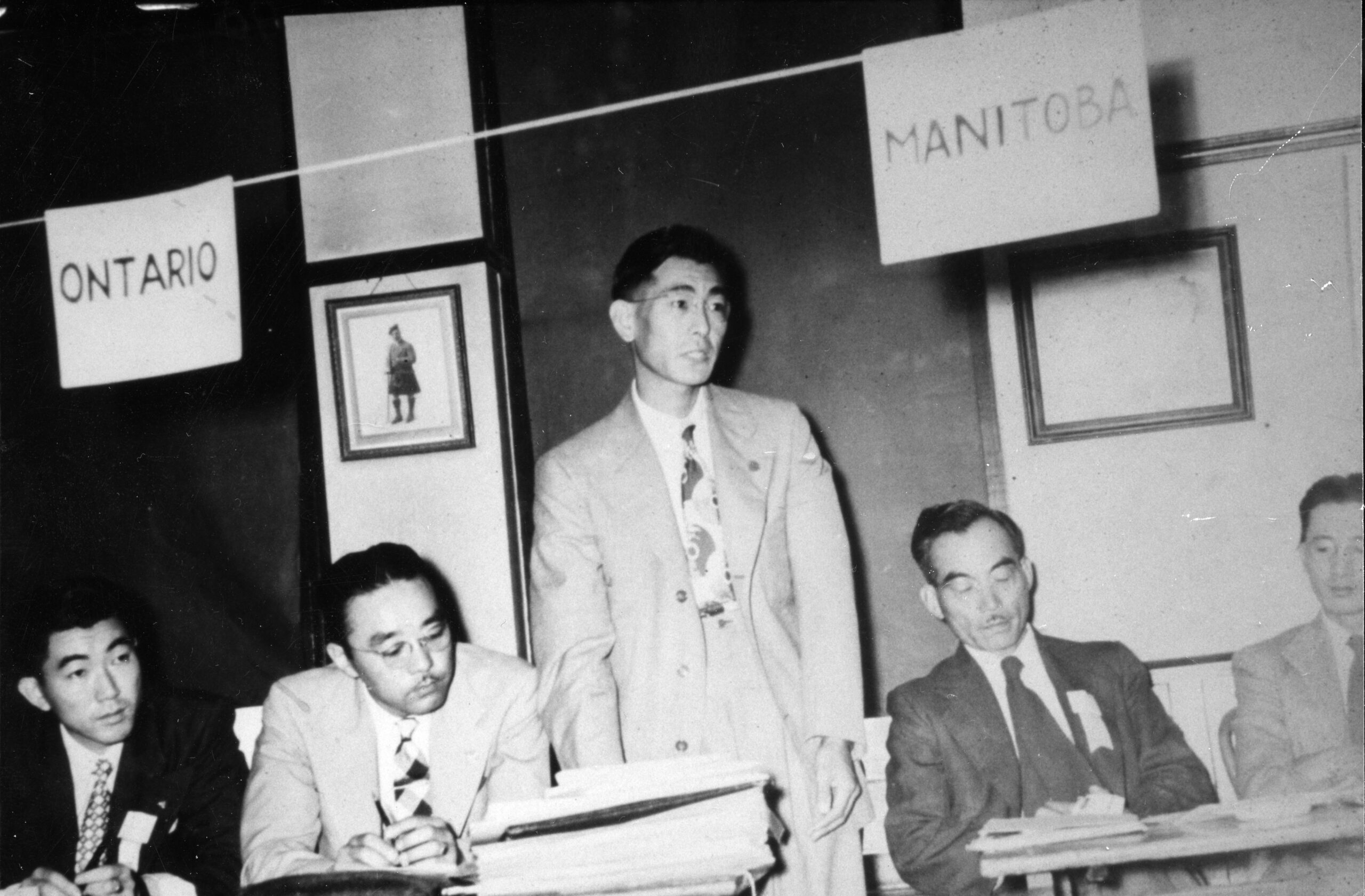 Four Asian men are seated at tables as one man stands and speaks. Signs above the tables read "Ontario" and "Manitoba."