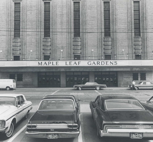 Exterior of an imposing building with a maruqee that reads "Maple Leaf Gardens". In the foreground are four parked cars.
