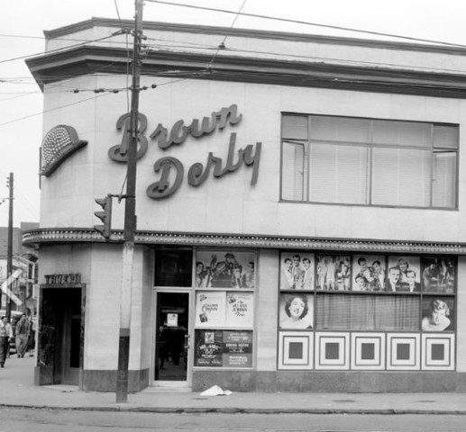 A black and white photo of a white building on the corner of a street, with a dark text as a sign that reads "The Brown Derby". There are images of people in the windows.