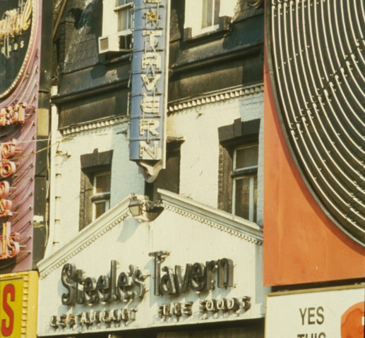 A colourful photograph depicting a bulb vertical sign on a white brick building with a black roof. The sign is blue and reads "Steeles Tavern".