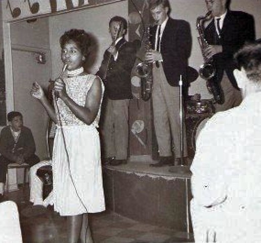 A black and white image of a female singer with dark skin singing into a handheld microphone, wearing a sequined light dress. Behind her on a stage stands a guitarist and two saxophone players, all men wearing suit jackets.