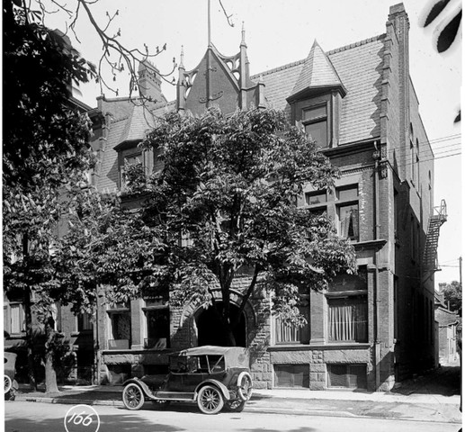 Black and white archival photo shows a three story building behind a large tree with an antique black automobile parked in front. The building is brick and has a staircase going up through a large arched entryway as well as distinctively triangular peaks on the roof.