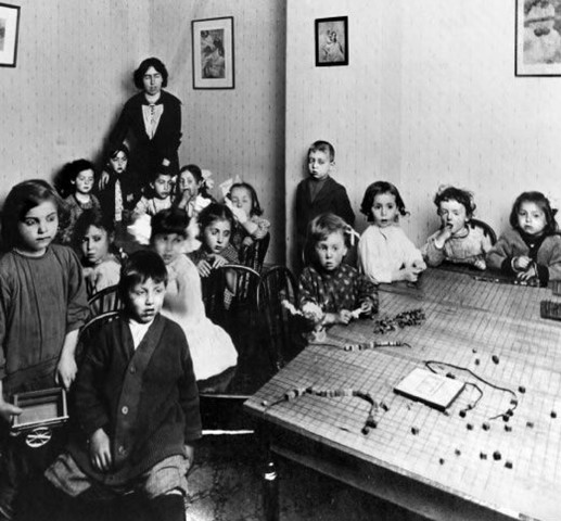 Black and white photograph of a room with children  facing the camera sitting in chairs around a table. The table has toys or games on it. In the back, behind the children, is a woman with dark hair standing facing the camera. Four images are hanging on the walls.
