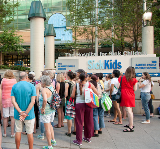 A crowd of people gather in a group in front of a large building with a sign that says, "The Hospital for Sick Children / SickKids" on a sunny day.