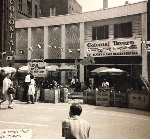 A black and white photograph of the front of Colonial Tavern.  People in 1960s style clothing walk along the street.