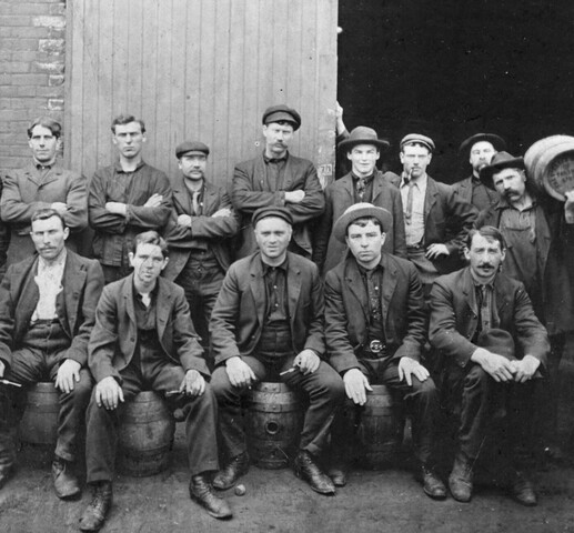 A black and white photograph of approximately 16 men seated and standing in front of an open warehouse door. The front row of men are seated on beer barrels. Behind them stand a row of men, most with their arms crossed. The two men on the far left and far right of the group are carrying beer barrels over their shoulders. Most of the men are wearing hats and are dressed in work clothes.