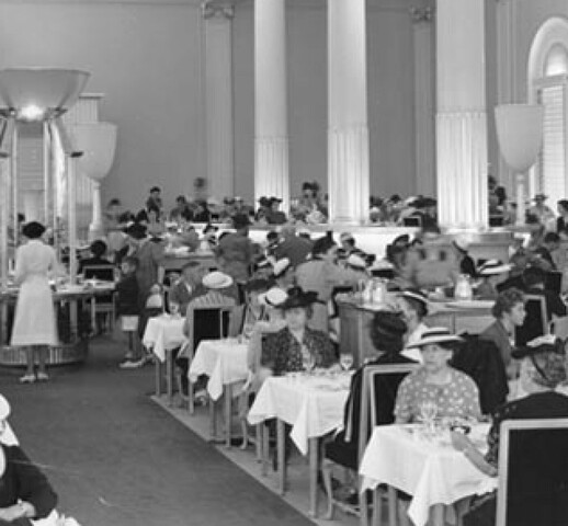 A black and white photograph of a bustling dining room, filled predominantly with women seated at tables, many of whom are wearing hats. The tables are covered with white tablecloths and the chairs are darkly upholstered. In the back of the room, a woman stands at a large circular table that is covered with dishes with food.