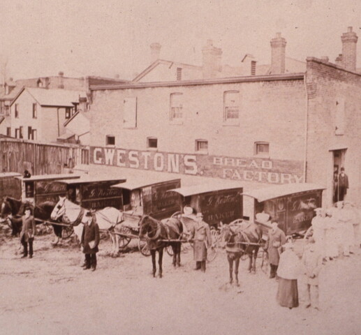 A faded photograph showing a bakery building labelled as "G. Weston's Bread Factory". In front of the building are six horse-drawn carriages, each labelled "G. Weston Baker". Men stand beside the horses. A group of individuals stand by the door of the building in baker's clothing.