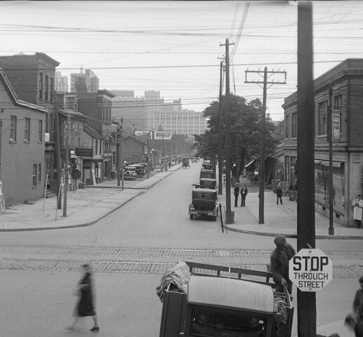 A black and white photograph of a street intersection. The streets are quiet and one pedestrian crosses the road. A stop sign reads: "Stop Through Street". Cars are parked on the right side of the street.