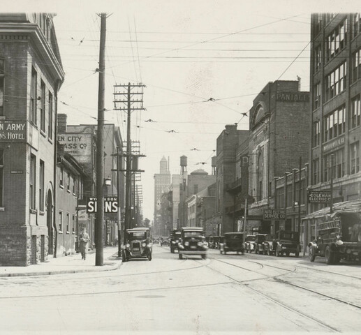 A black and white photograph of a street scene. Several brick buildings on either side of the road. Old cars from the 1920s can also be seen driving and parked on the road.