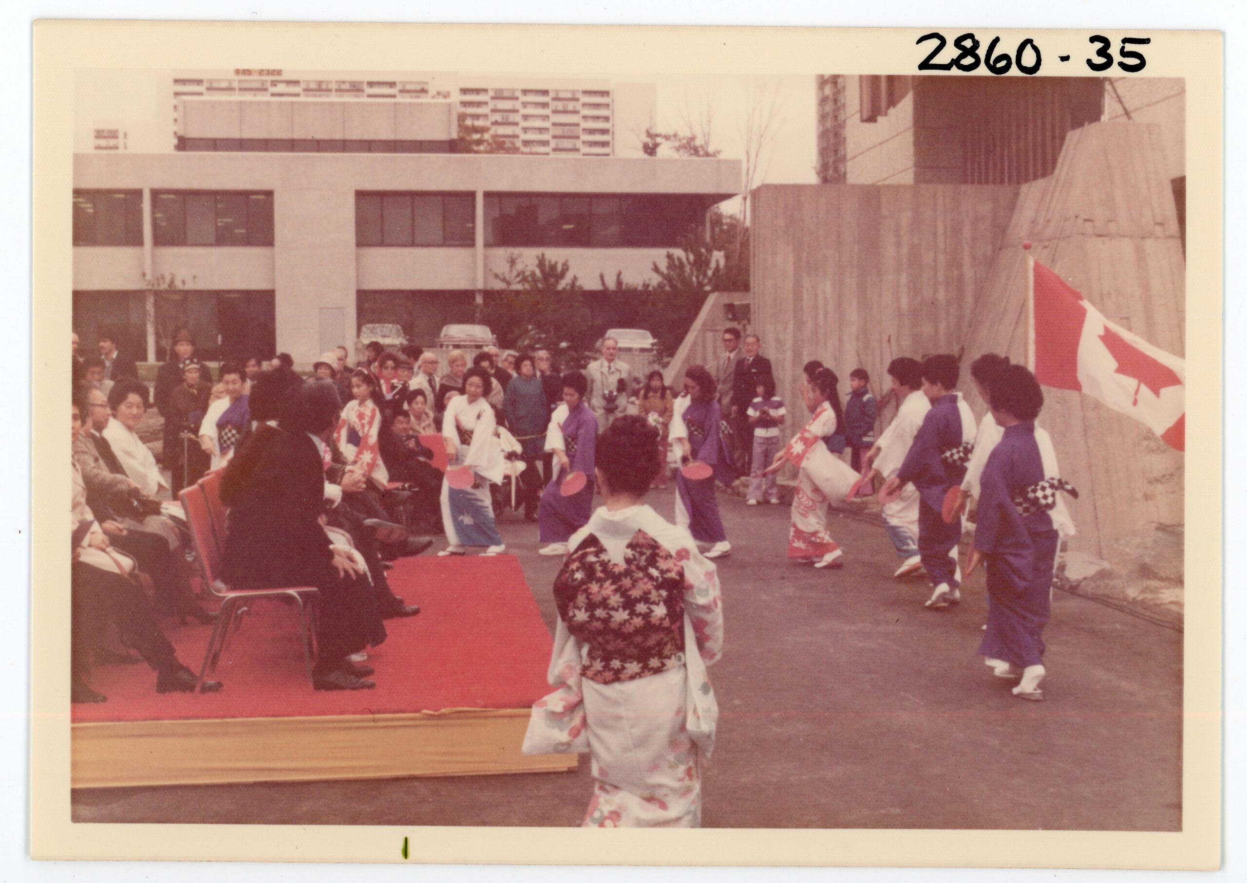 A group of dancers wearing kimonos performs around a raised platform where guests are seated. A Canadian flag is at right.