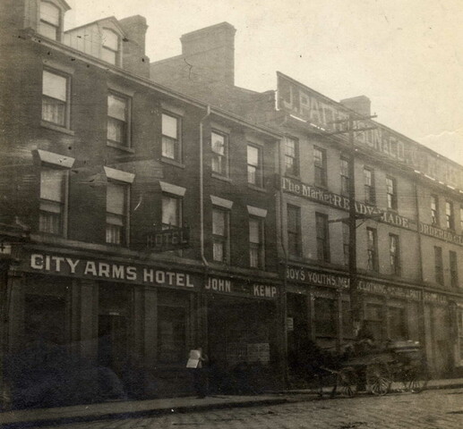 Black and white photo of a cobblestone street with buildings alongside it. One building's sign reads, "City Arm Hotel" and beside that, "John Kemp". The sign on the building beside this reads, "Patterson & Co." and various other shop signs are visible on the same building. A horse-drawn carriage is in front of the shops on the road.