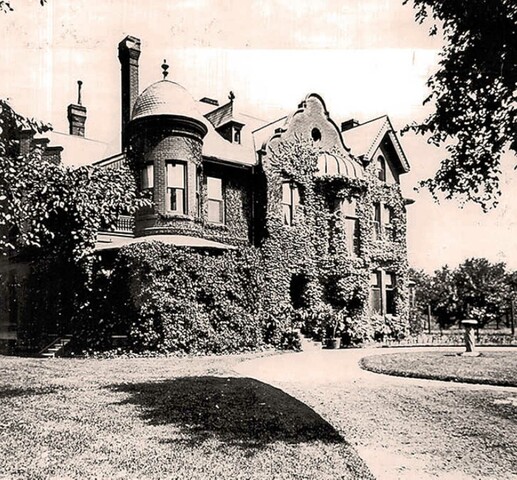 Black and white photo of a large house with many vines covering it. A circular driveway is in front of the house and trees are visible in the distance. In the middle of the driveway is a statue or fountain.