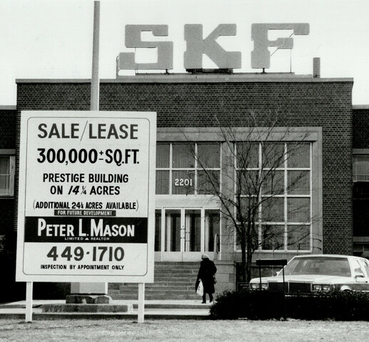 Black and white photo of a large building with the letters "SKF" on top of it. There is a car parked in front of it and there is a large sign that reads, "Sale/Lease / 300,000 SQ FT. / Prestige Building / ON/14 1/4 Acres / Additional 24 1/4 acres available /  for future development / Peter L. Mason / limited realtor / 449-1710 / inspection by appointment only."