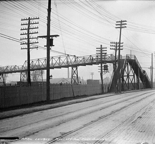 Black and white photograph of a large steel bridge in the distance and a road in the foreground. Telephone poles run alongside the road.