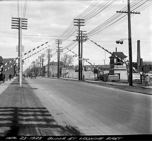 Black and white photograph of a city road with many telephone poles and crossing barriers alongside it. The crossing barriers are striped and are raised in the air. In the background are industrial buildings.