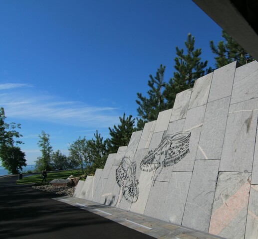 Photo of a walkway in a park. The photo is taken from under an underpass and on the grey stone wall of the underpass is an engraving of a pair of moccasins. Trees are visible in the distance.