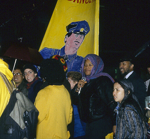 Leleti Tamu, member of the Black Women’s Collective, is surrounded by others at the Sophia Cook protest. There is a yellow banner in the background with an illustration of a police officer on it.