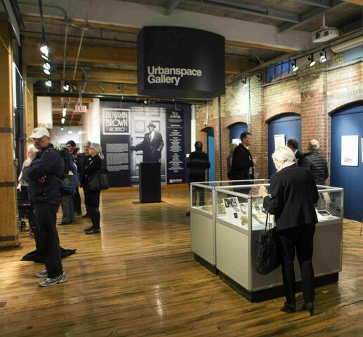 Image of an architect exhibition. There are people walking around looking at art work and displays. There are brick walls surrounding the room with blue painted garage doors on the right side of the room, illuminated with bright lights on top. There is a big, black sign hanging from the ceiling that reads "Urbanspace Gallery."