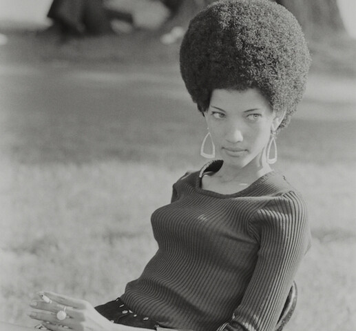 Black and white photograph of a woman sitting in a chair outside. She has her hair in a tall afro and is wearing large drop earrings. She is wearing a long-sleeved ribbed knit sweater and plaid shorts. She is holding a cigarette in her left hand. Behind her out of focus is a large tree with people sitting against it.