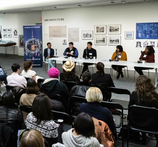 Room filled with lots of people seated listening to other people speak, seated in the front of the room. There are posters and photographs framed along the walls and tables with glass tops lined against the walls around the room.