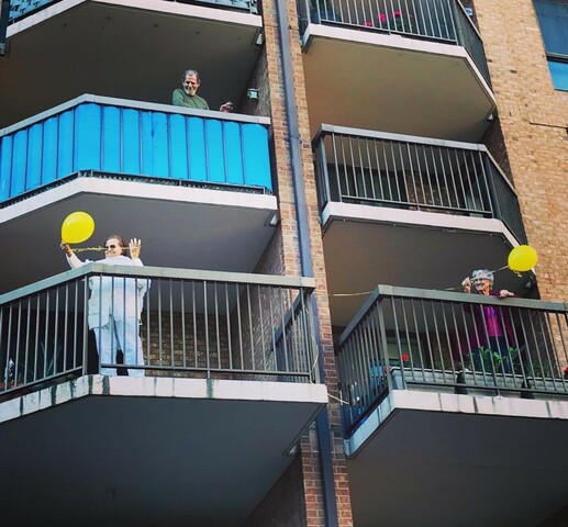 Photograph of apartment building balconies. On the two lowest balconies are two women who are holding yellow balloons. On the middle left balcony is a man looking down at the lower balcony.