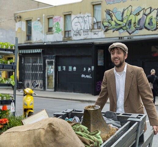 Photograph of a man pushing a large wooden cart. In the cart is a large potato sack. He is dressed in a white collared shirt, a brown suit jacket, and a brown newsboy hat. Behind him is a couple pushing a stroller, a yellow moped and a black building with graffiti on the top most wall.