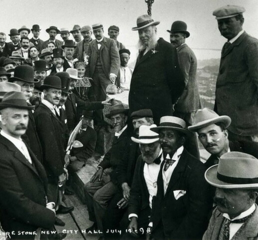A group of men are gathered on narrow wooden walkway above a city skyline. Most are wearing hats and face the camera.