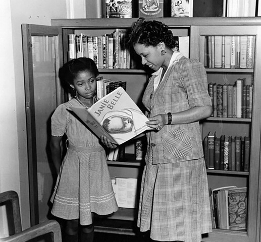 A woman, standing, holds open a book. To her left, a young girl looks at the book the woman holds. Behind them is a bookshelf filled with books.