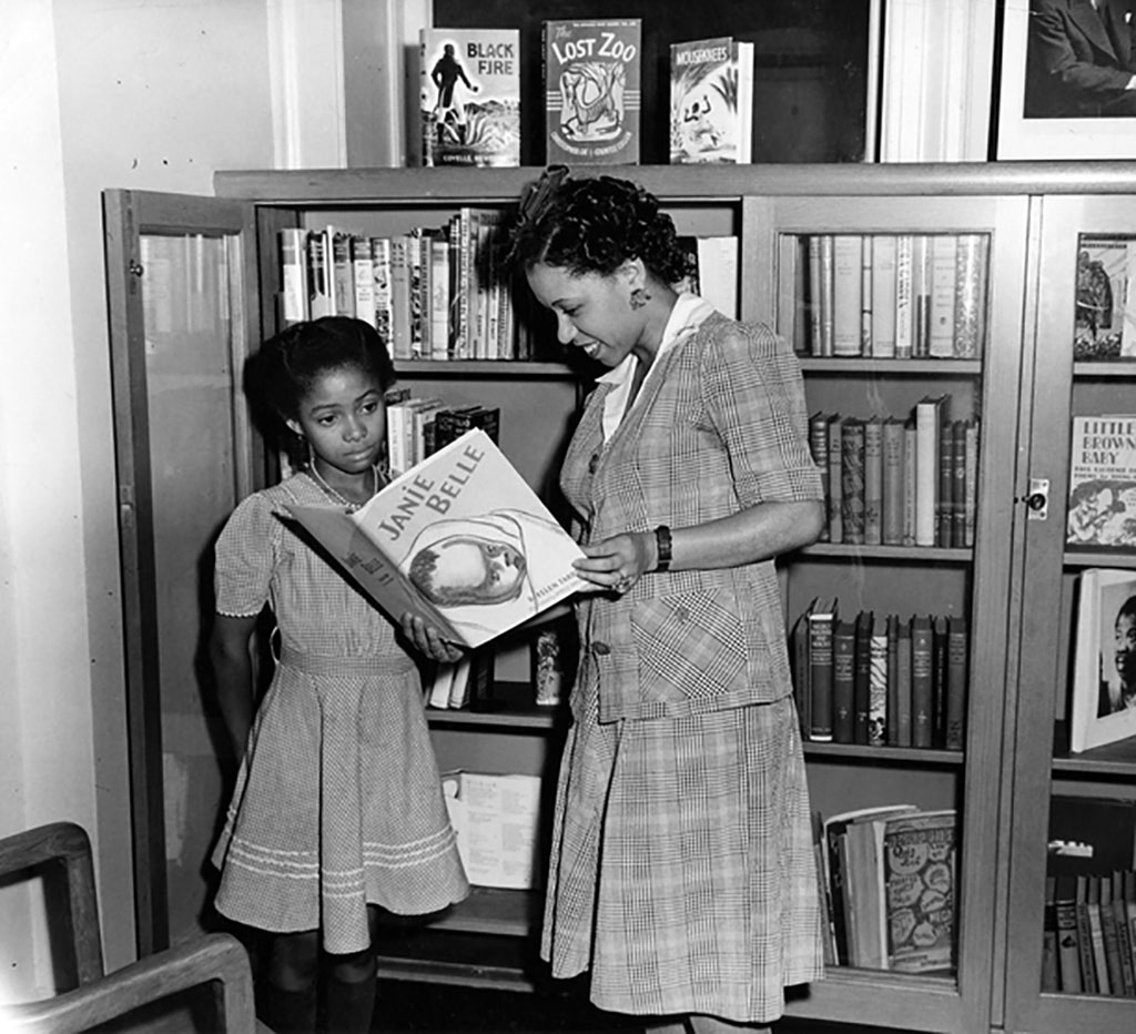 A woman, standing, holds open a book. To her left, a young girl looks at the book the woman holds. Behind them is a bookshelf filled with books.