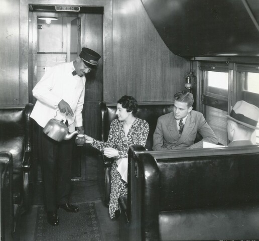 A man in a white uniform pours tea to a woman seated in a train car booth. A man seated next to her looks on.
