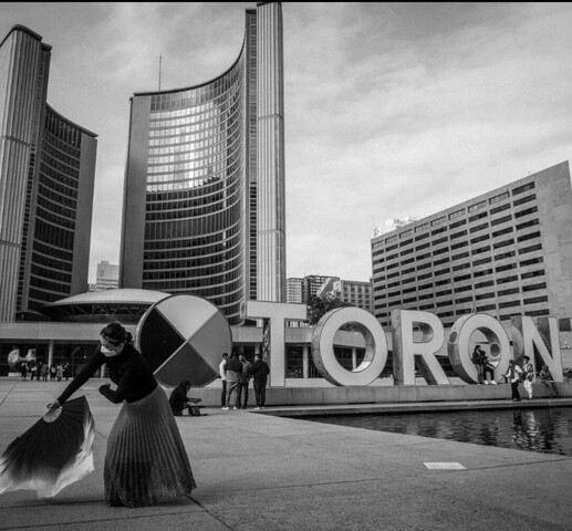 A black and image showing a large sign with circular graphic image with four quadrants and the words "TORONTO" on the right hand side of the image. There is a person sitting in the letter "O" structure behind a group of people standing near the sign along a pool or pond of water. A woman in a dark top and long skirt is bent over, holding a large hand fan on the left hand foreground of the image. There are tall, multi-story buildings in the background.