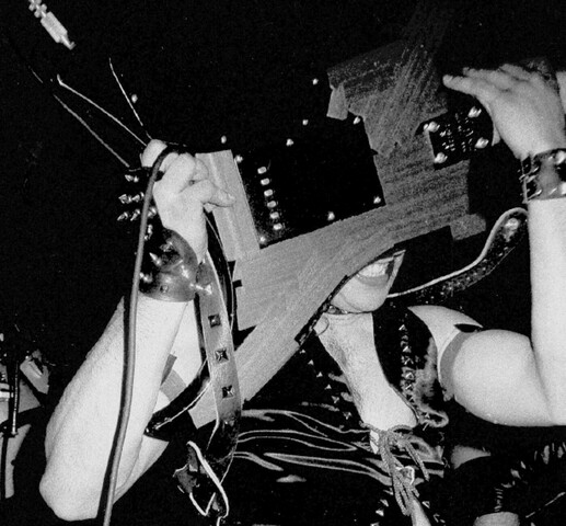 A black and white photo of a man holding an electric guitar in front of his face. He is wearing a leather outfit. The only part of his face that is visible under the guitar is his smiling mouth.