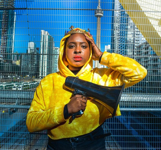 A woman in a yellow camouflage hoodie wearing a gold crowing with red jewels and red lipstick holds a megaphone on a bridge with the CN tower and multiple skyscrapers in the background.