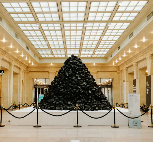 A pile of hundreds of black conductor caps sits on a white pedestal in the middle of a large beige room with potlights and a skylight.