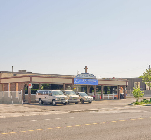 Street view image of single story building with a curved signboard and cross on the front façade. There are 3 white vans parked on the left in front of the building and small trees on the right side.