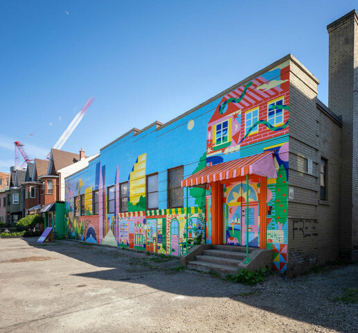 Colour photo of a block of buildings. The most prominent building has a large colourful mural painted on the front facade with a red and white awning over the door. There are multi story buildings  in the background with construction cranes.