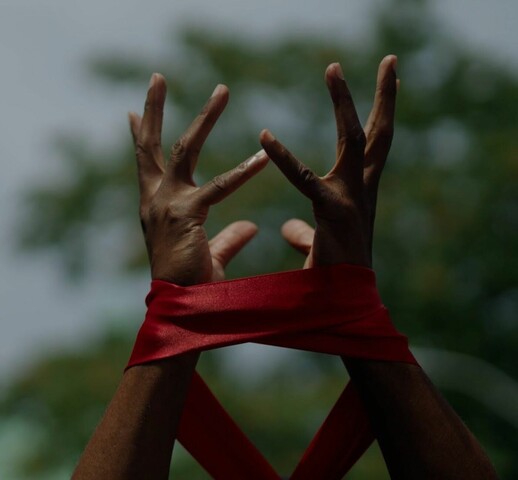 Colour photo of two hands raised towards the sky with wrists loosely wrapped in a dark red fabric.