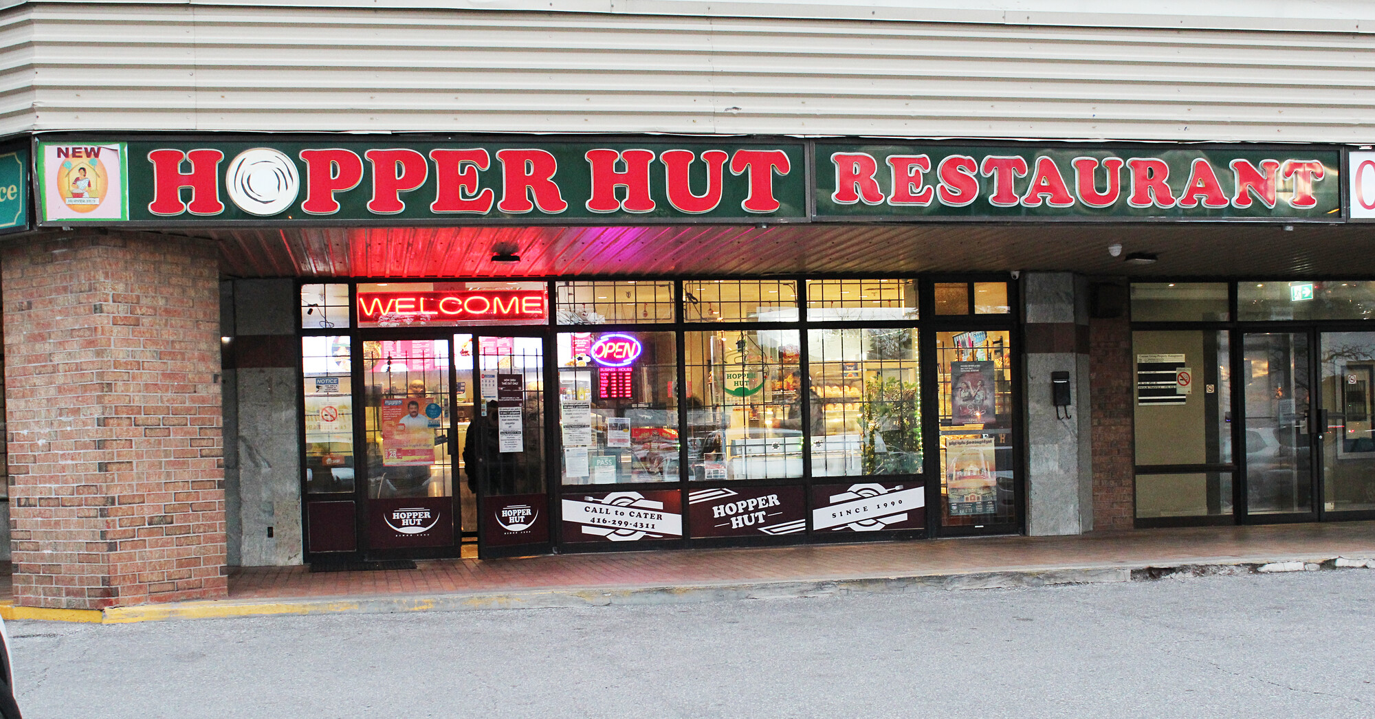 Exterior of a restaurant. Over the door, the words "Hopper Hut Restaurant" in red letters appear over a dark green background. Through the restaurant windows, a takeout counter with customers gathered around is visible.