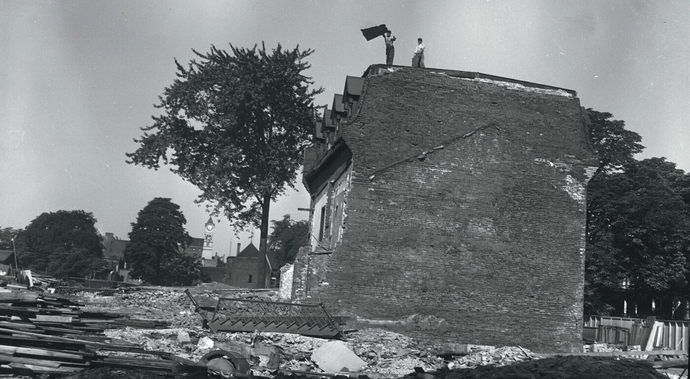 Image of a row house being demolished. Two men are on the roof and a staircase is visible lying on the ground.