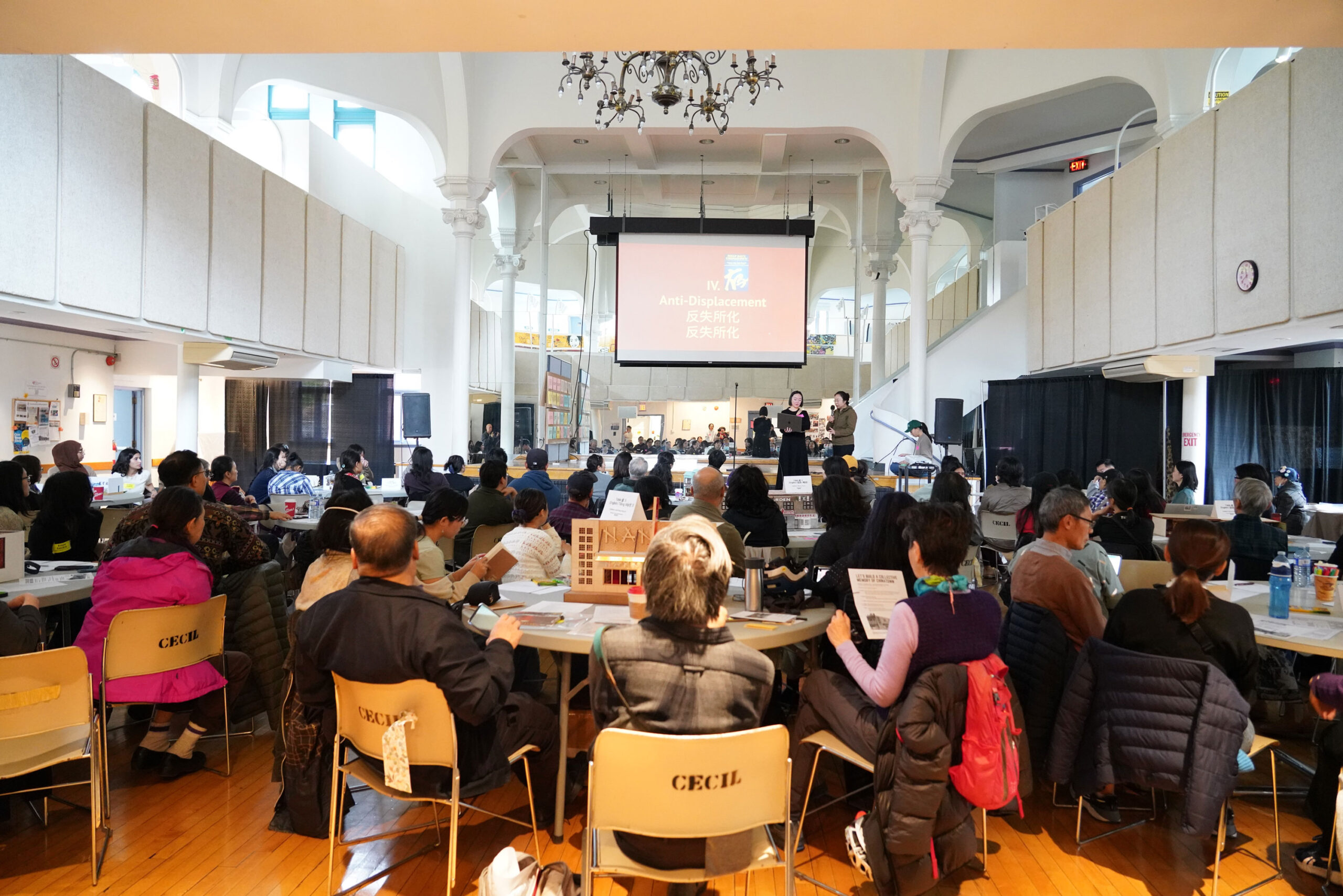 A crowd of people in a conference hall sitting on round banquet tables watching a presentation on the main stage.