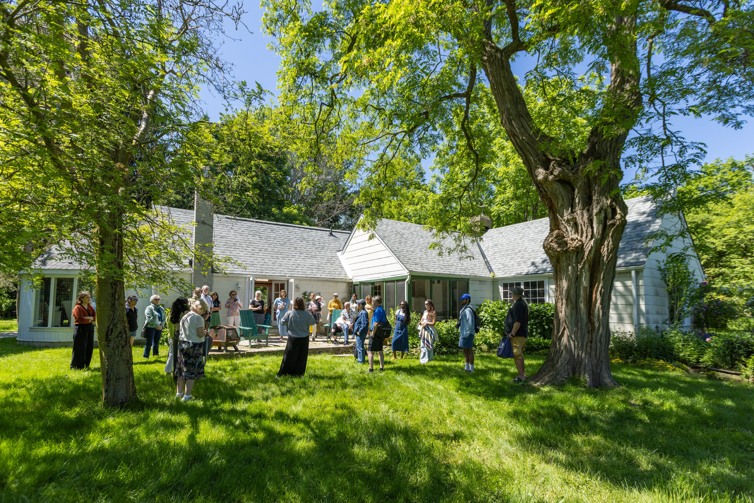 A crowd of people listening to a speaker. All are standing on the lawn of a white-walled and grey-roofed cottage.