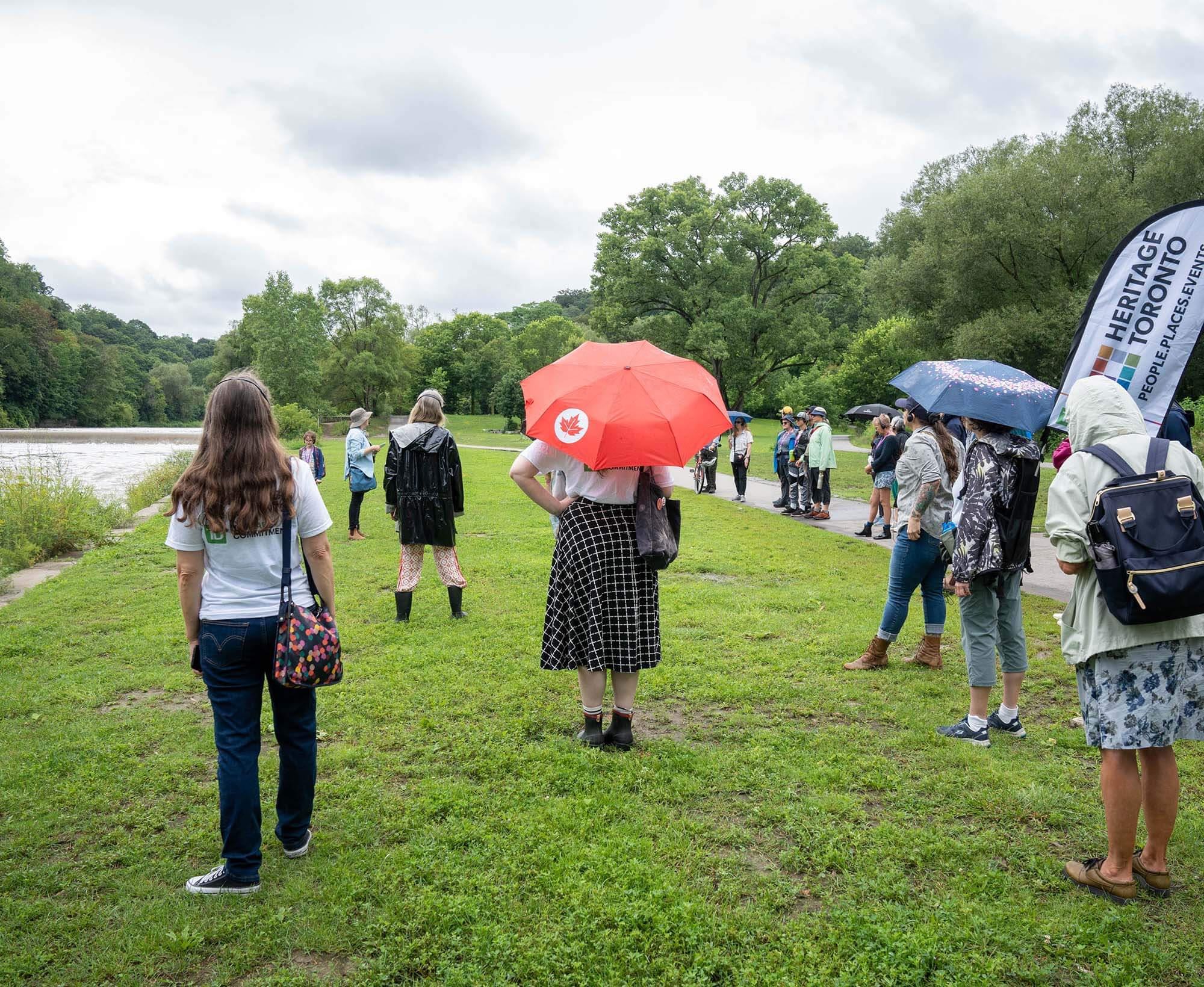 Group of people stand on grass next to a river in a park-like setting.