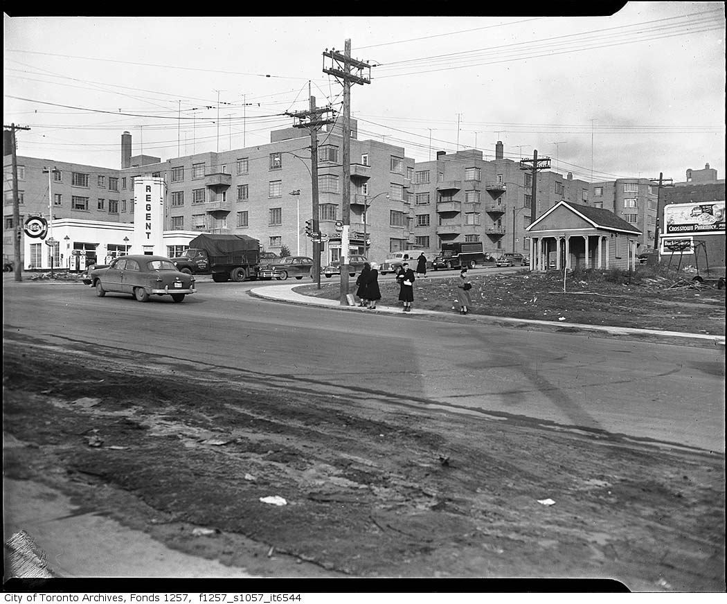 A black and white image of a street corner with dirt roads, people walking on the street, visible electric poles and low-rise apartments in the background.