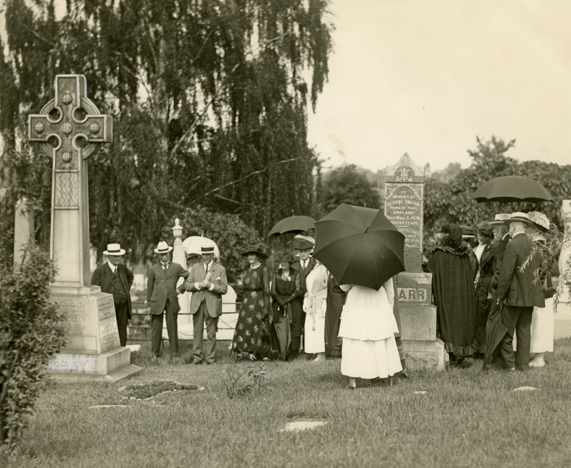 Black and white image of men and women surrounding a grave approximately 12 feet tall topped with a Celtic cross. The people are wearing black and white clothing and some are carrying black umbrellas. Other graves are visible around them.